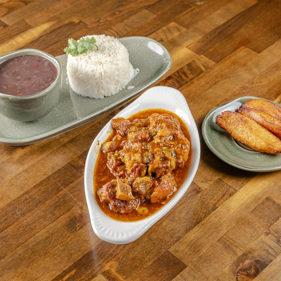 Rice, bean soup, saucy stew, and fried plantains arranged on plates over a wooden restaurant table.