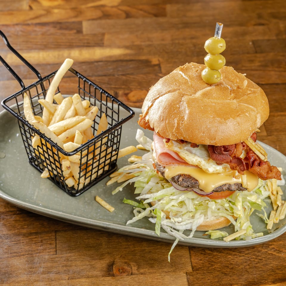 Loaded bacon cheeseburger with fried egg and olives, served with French fries in a basket on a wooden table.
