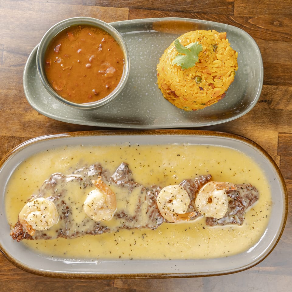 Creamy shrimp-topped steak served with spiced yellow rice and a bowl of reddish lentil sauce on wooden table.