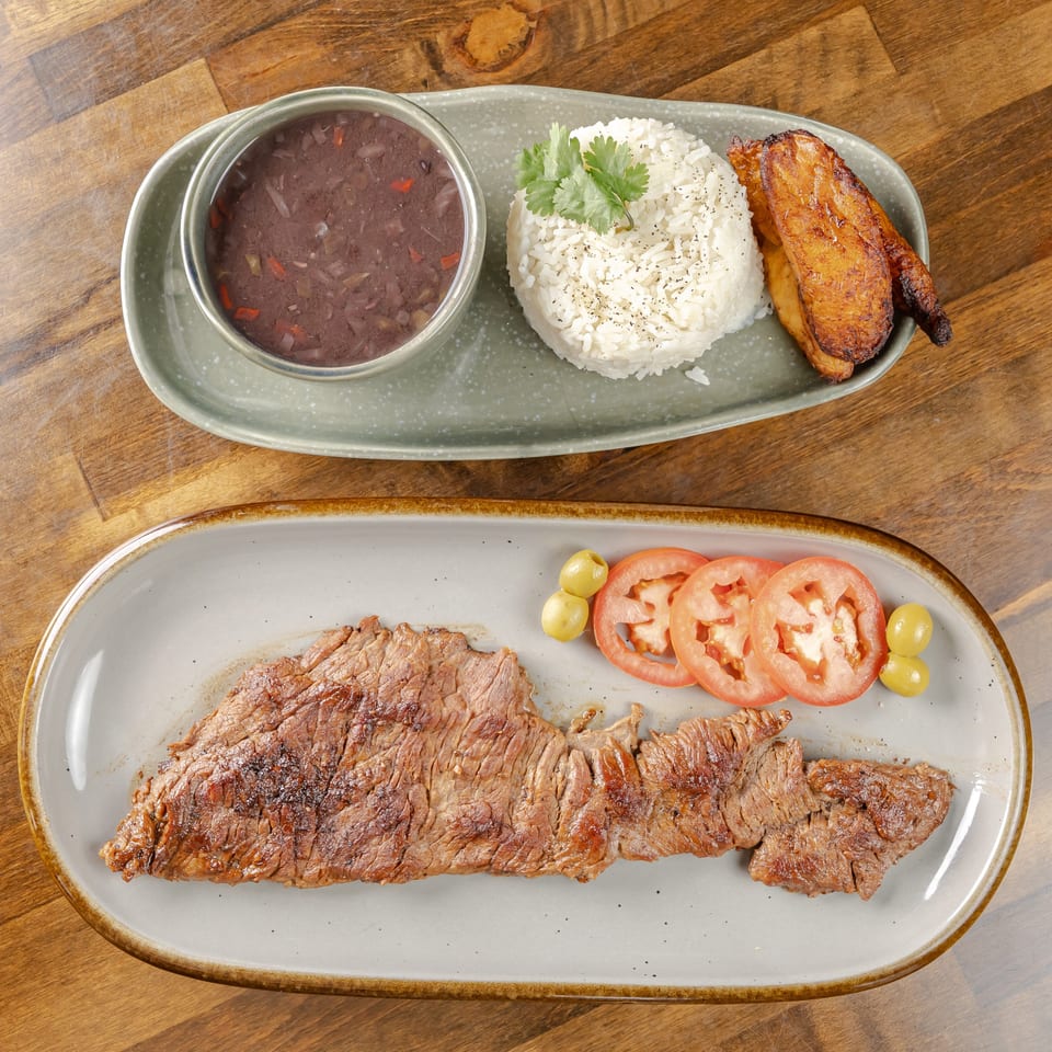 Grilled steak with sliced tomatoes and olives, alongside rice, black beans, fried plantains, and cilantro on plates.