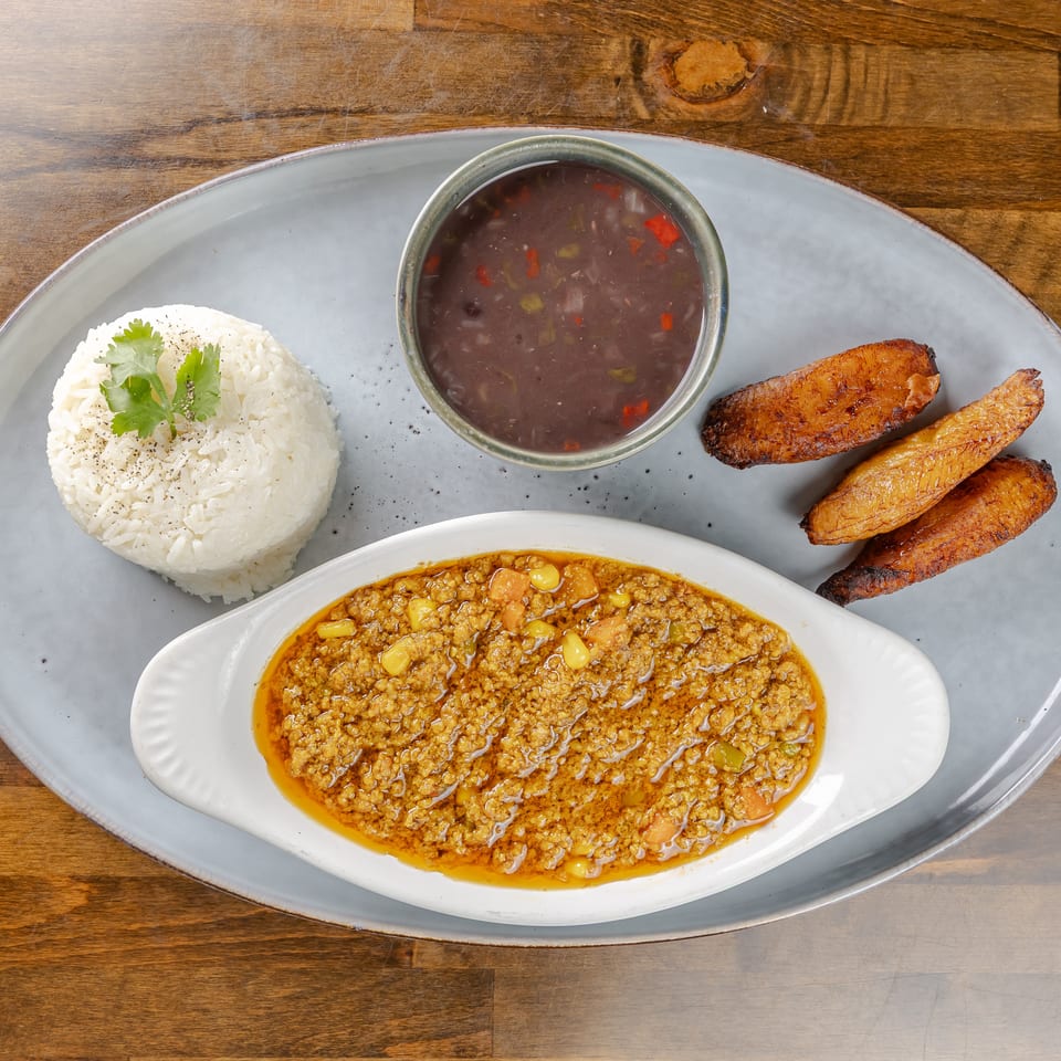 Plated Latin American meal with white rice, stewed beans, ground meat in sauce, and fried plantains on a gray platter.