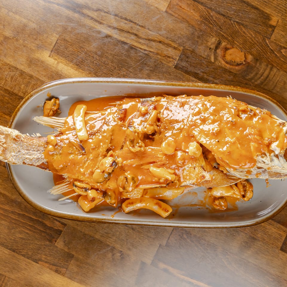 Whole fried fish covered in creamy orange sauce and seafood pieces, served on a platter atop a wooden table.