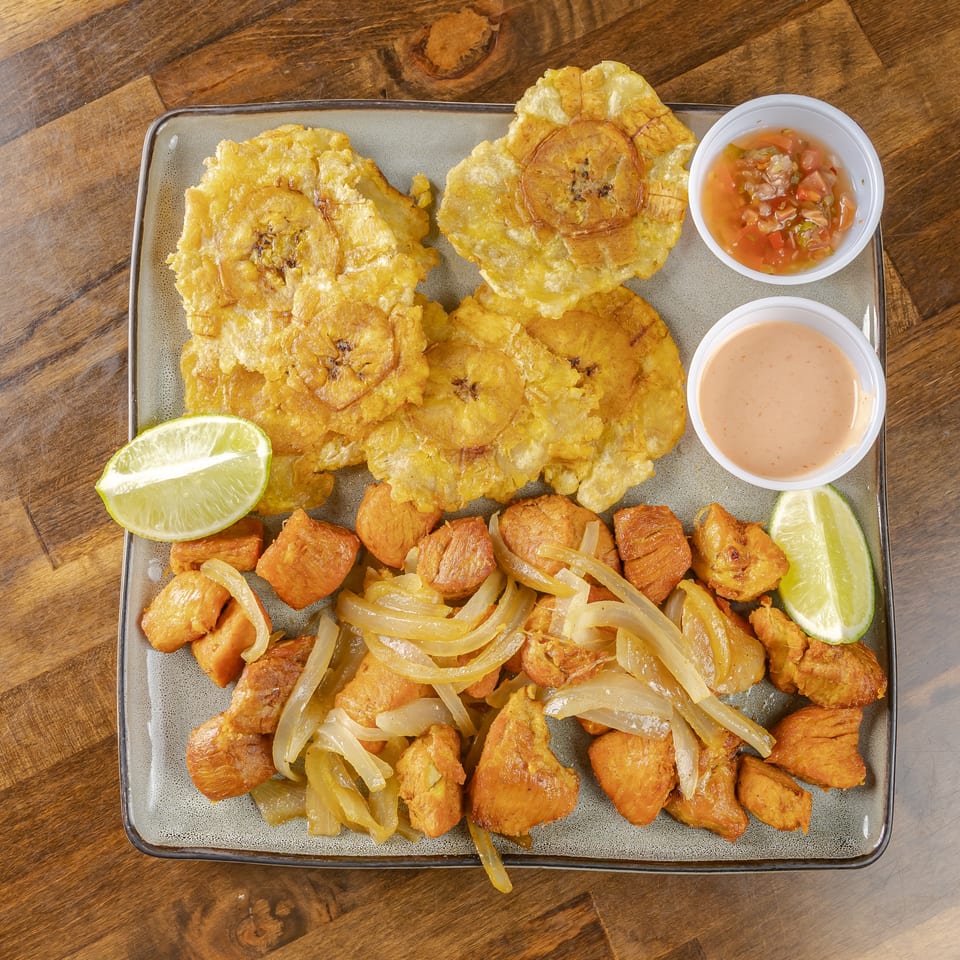 Platter of fried plantains, golden fried pieces, lime wedges, and two dipping sauces on a wooden table.
